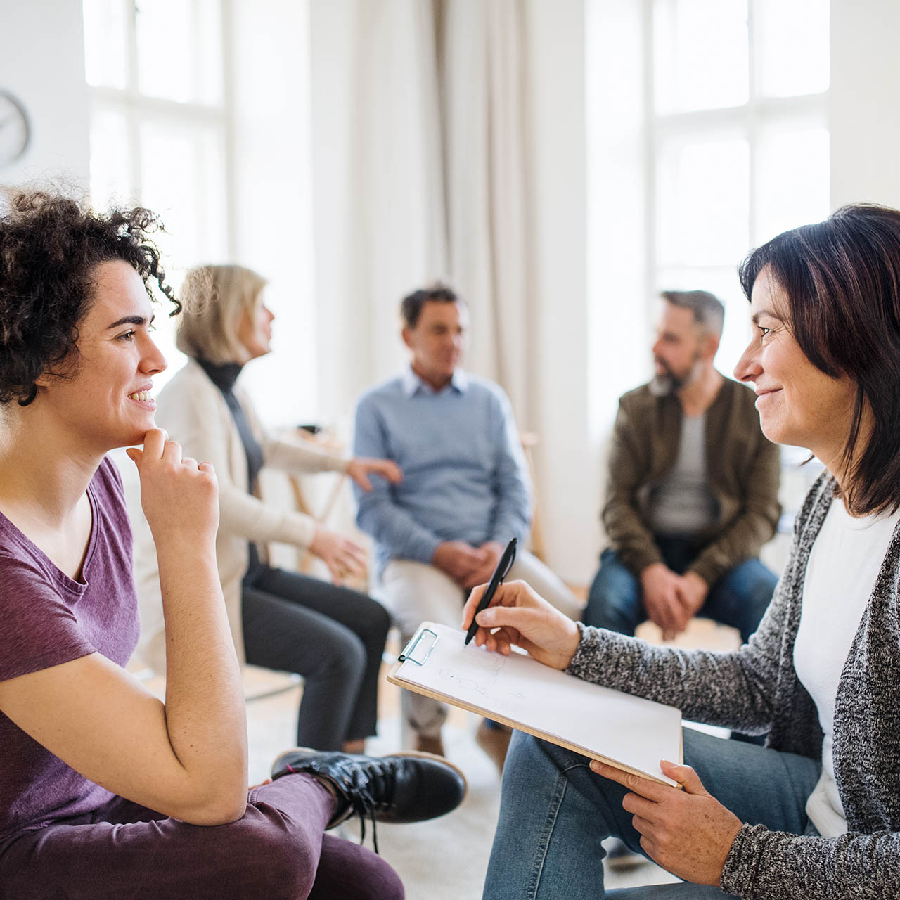 Senior counsellor with clipboard talking to a woman during group therapy.