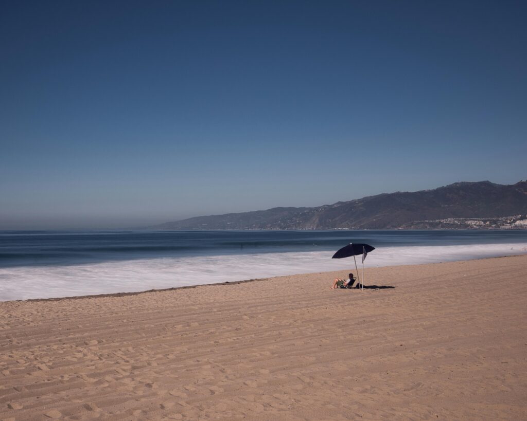 Southern California Beach at noon with one person sitting under an umbrella on the sand.