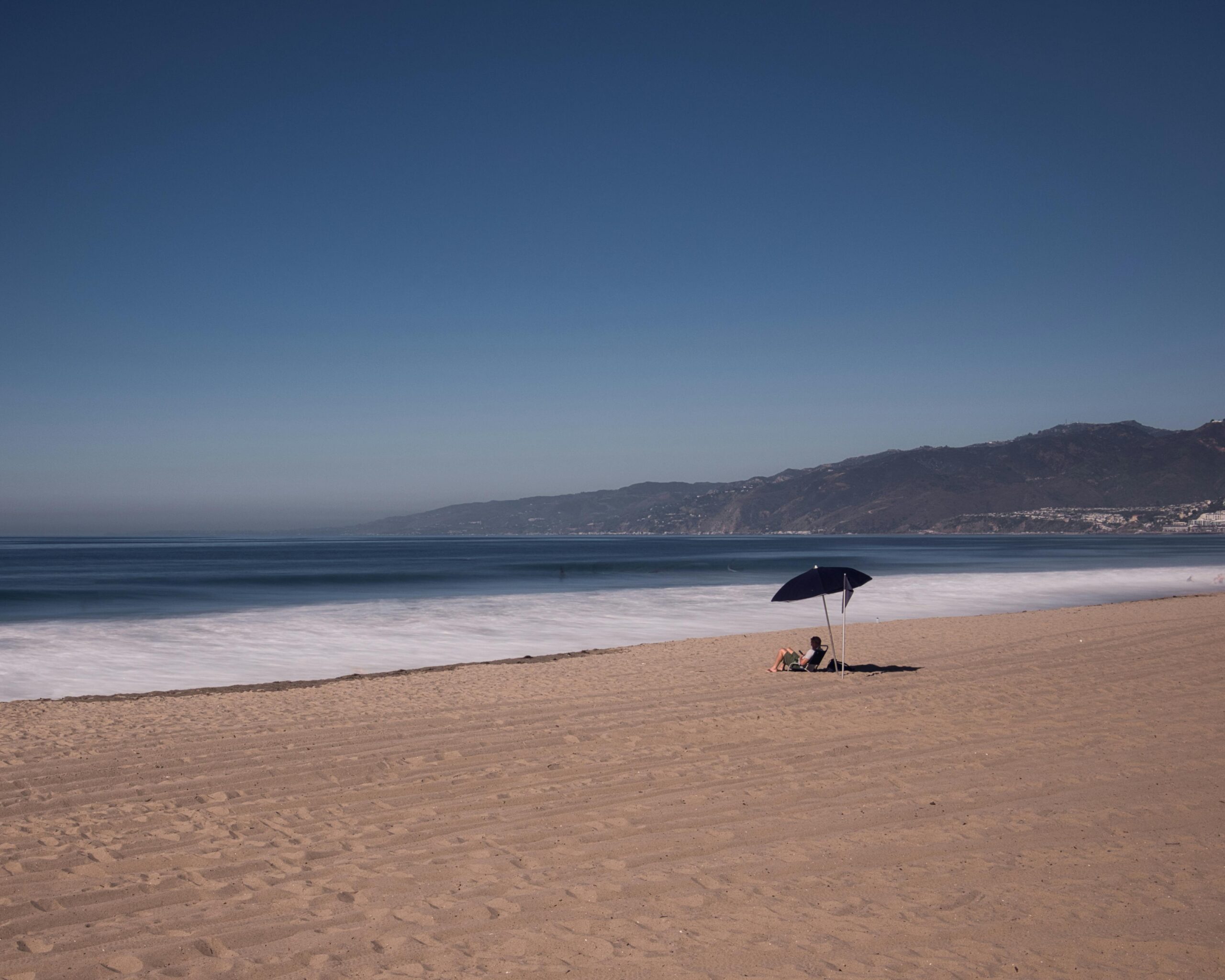 Southern California Beach at noon with one person sitting under an umbrella on the sand.